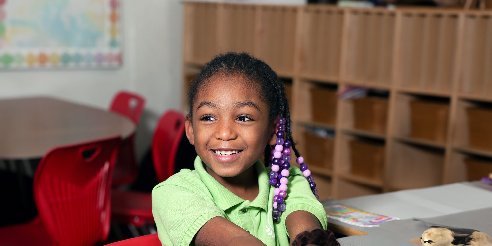 Smiling student with braided hair and purple beads in a classroom.